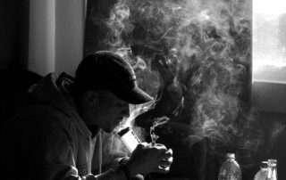 Pessoa sentada em ambiente fechado, com fumaça ao redor, ilustrando sinais associados à dependência química.